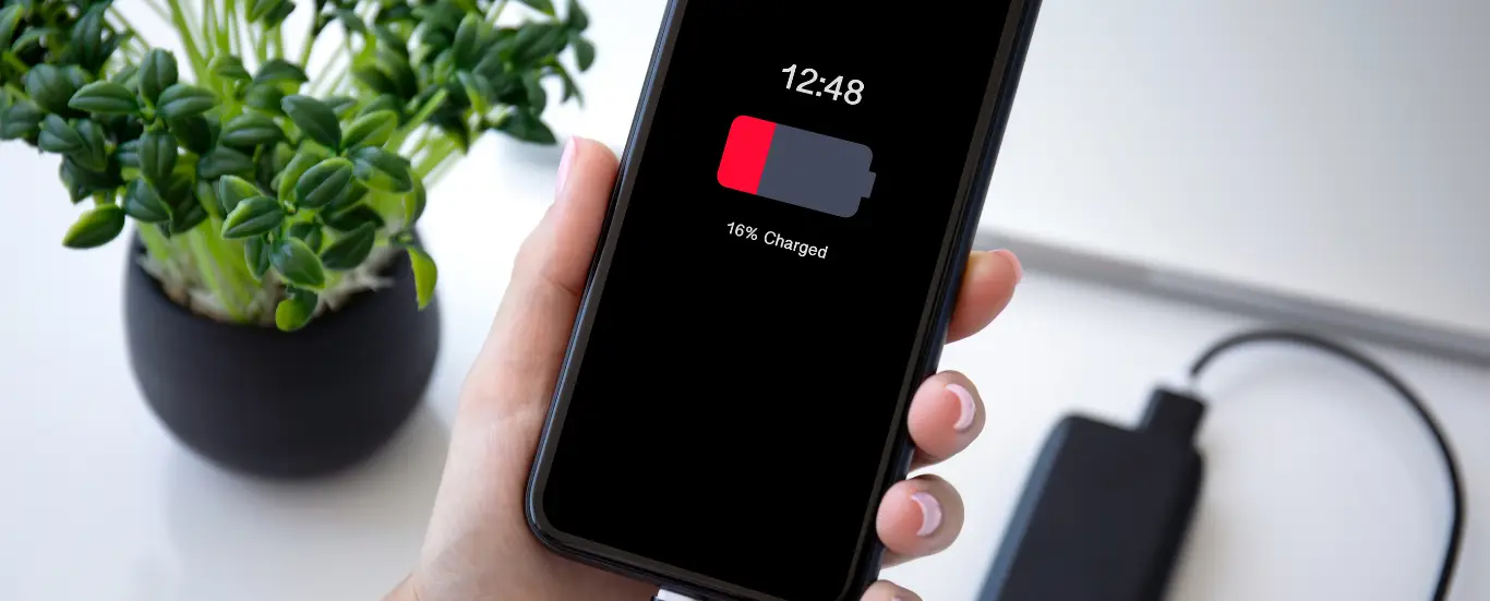 Hand holding a smartphone showing a low battery charging screen next to a charger on a white desk.